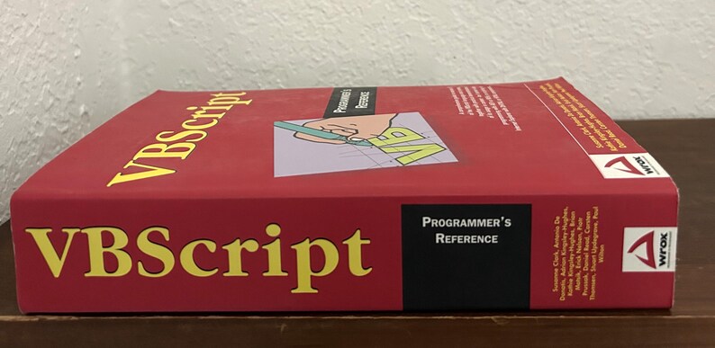 May include: A red VBScript Programmer's Reference book with yellow text on the cover. The cover features a graphic of a hand holding a green pen over a grid with the letters VB. The Wrox logo is visible on the side and the spine of the book.