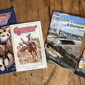 May include: Four vintage Calgary Stampede event brochures and guides. The brochures feature images of rodeo events, cityscapes, and attractions. The color palette includes blue, red, white, and brown. The brochures are arranged on a wooden surface.