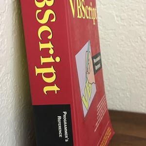 May include: A thick red reference book titled VBScript Programmer's Reference with yellow text on the spine and front cover. The cover features a graphic of a hand writing on a page, the Wrox logo, and a list of authors including Susanne Clark.