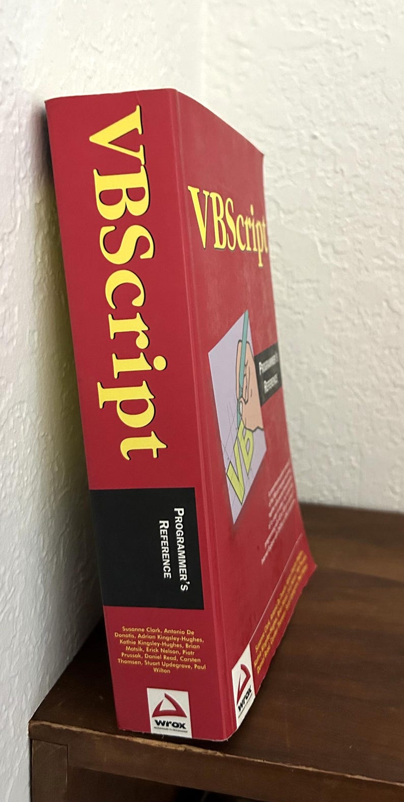 May include: A thick red reference book titled VBScript Programmer's Reference with yellow text on the spine and front cover. The cover features a graphic of a hand writing on a page, the Wrox logo, and a list of authors including Susanne Clark.