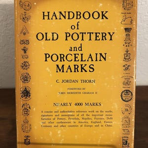 May include: A vintage book titled "Handbook of Old Pottery and Porcelain Marks" with a yellow cover and black text. The book features various pottery marks around the border. The book is held by a metal stand.