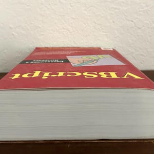 May include: A thick paperback book titled VBScript Programmer's Reference with a red cover. The front cover features yellow text and a small graphic illustration of a hand holding a green pen. The book is positioned horizontally with white pages.