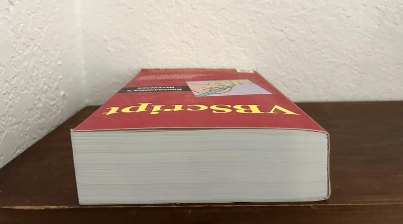 May include: A thick paperback book titled VBScript Programmer's Reference with a red cover. The front cover features yellow text and a small graphic illustration of a hand holding a green pen. The book is positioned horizontally with white pages.