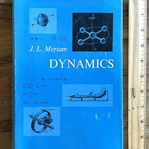 May include: A blue textbook titled Dynamics by J.L. Meriam, featuring scientific diagrams of a gyroscope, molecular structure, aircraft, and ring gear, alongside mathematical equations, positioned next to a wooden ruler measuring 11 inches in length.