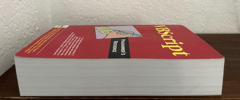 May include: A thick paperback book titled VBScript Programmer's Reference with a red cover. The cover features a graphic of a hand holding a green pen over the letters VB. The text is printed in yellow and white against a red and black background.