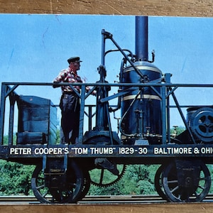 May include: Vintage postcard featuring Peter Cooper's "Tom Thumb" steam locomotive from 1829-30, Baltimore & Ohio R.R. The black and blue locomotive is on a flatbed with a man standing on it. The background shows a blue sky and green trees.