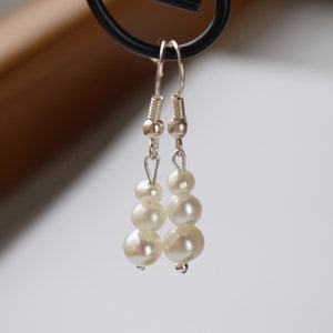 May include: A pair of silver dangle earrings with three white pearls on each earring.