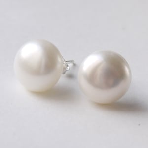 Op de afbeelding: Twee witte parelknopjes. De parels zijn rond en hebben een glanzende glans. De oorbelpalen zijn zilver. De oorbellen staan tegen een witte achtergrond.