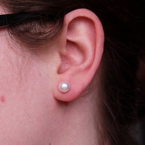 May include: A close-up of a person's ear with a white pearl earring. The earring is a simple stud style.