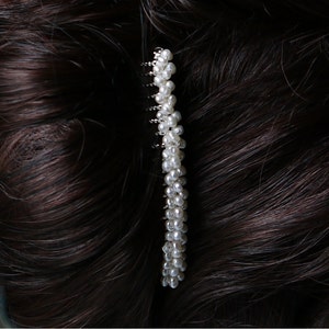 Puede incluir: Un peine para el cabello de perlas blancas con una base de metal plateado. El peine está hecho de una fila de pequeñas perlas blancas.