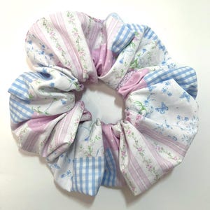 May include: A large, handmade scrunchie with a patchwork design. The fabric features a mix of patterns, including pink stripes, blue gingham, and floral prints in pink and blue. The scrunchie is designed to hold hair.