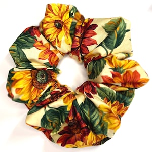May include: A fabric hair scrunchie featuring a repeating pattern of yellow and deep red sunflowers with dark green leaves on a cream-colored background. The accessory has a gathered, ruffled texture and a circular shape with a central opening.