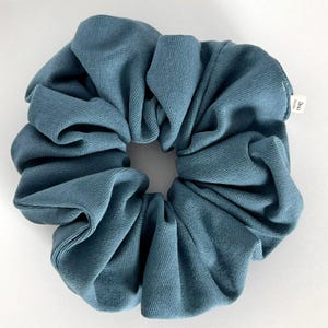 May include: A large, teal-colored scrunchie made of a textured fabric. The scrunchie is circular with a gathered design, and a small white tag is visible. This hair accessory is designed to hold hair in a ponytail or bun.