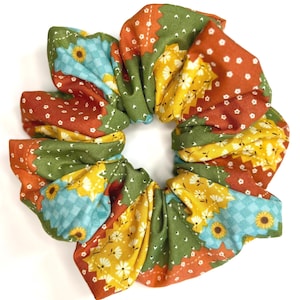 May include: A fabric hair scrunchie featuring a patchwork design with various floral and geometric patterns in shades of olive green, burnt orange, mustard yellow, and light blue, with small white flower accents throughout the textile.