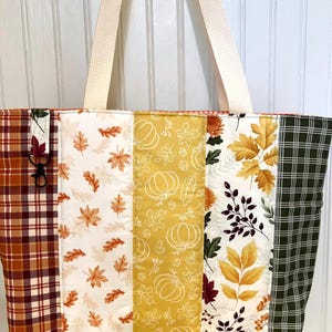 May include: A quilted tote bag featuring vertical panels of autumn-themed fabric. The patterns include orange and brown plaid, white with scattered maple leaves, yellow with white pumpkin outlines, white with fall foliage, and dark green with a white grid.