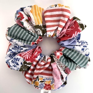 May include: A large, colorful scrunchie made from various patterned fabrics. The scrunchie features red and white stripes, floral prints, and other designs. A small tag with the text "Olivia's" is visible.