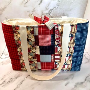 May include: A quilted tote bag featuring a patchwork design of red, white, and blue fabrics. The exterior displays various patterns including gingham, plaid, and star motifs. It has two cream-colored fabric handles and a matching fabric tie closure.