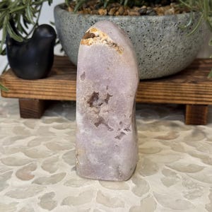 May include: A pale purple crystal geode with a textured surface. The crystal has a rounded shape with a cavity in the centre. The geode is displayed on a wooden shelf with a black bird figurine and a potted plant in the background.