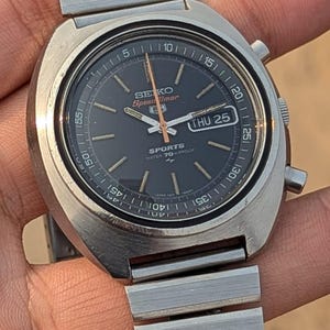 May include: A vintage Seiko Speedtimer watch with a black dial, silver hands, and a date display. The watch has a stainless steel case and bracelet. The dial features the text "SEIKO Speedtimer SPORTS WATER 70 PROOF".