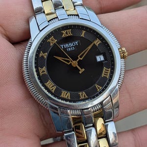May include: A Tissot 1853 wristwatch with a black face, gold hands, and Roman numeral hour markers. The watch has a silver and gold-tone stainless steel band. The date is displayed at the 3 o'clock position.
