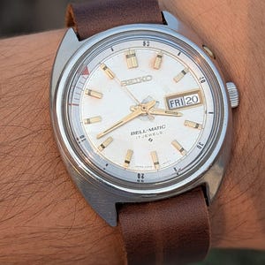 Puede incluir: Un reloj de pulsera Seiko Bell-Matic vintage con esfera plateada, agujas e indicadores dorados y correa de cuero marrón. La esfera del reloj muestra el día y la fecha. El reloj tiene una caja de acero inoxidable.