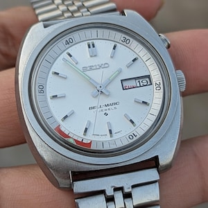 May include: A vintage Seiko Bell-Matic watch with a silver case and strap. The white watch face features black and silver markers, and the date is visible. The watch has a distinctive design with a side button and a red and green accent.