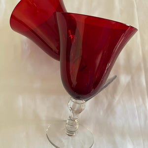 May include: Two ruby red wine glasses with a clear stem and base. The glasses have a tulip-shaped bowl and are made of glass. The glasses are set against a white background.