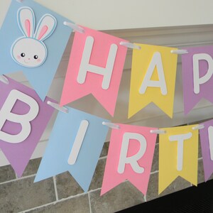 Puede incluir: Una pancarta de colores pastel con las palabras "Happy Birthday" en letras blancas. La pancarta tiene un conejito con orejas rosas en la primera bandera.
