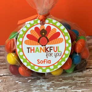 Op de afbeelding: Een ronde groene en witte tag met een kalkoenillustratie en de tekst "THANKFUL for you! Sofia". De tag is bevestigd aan een doorzichtige plastic zak gevuld met kleurrijke snoepjes.