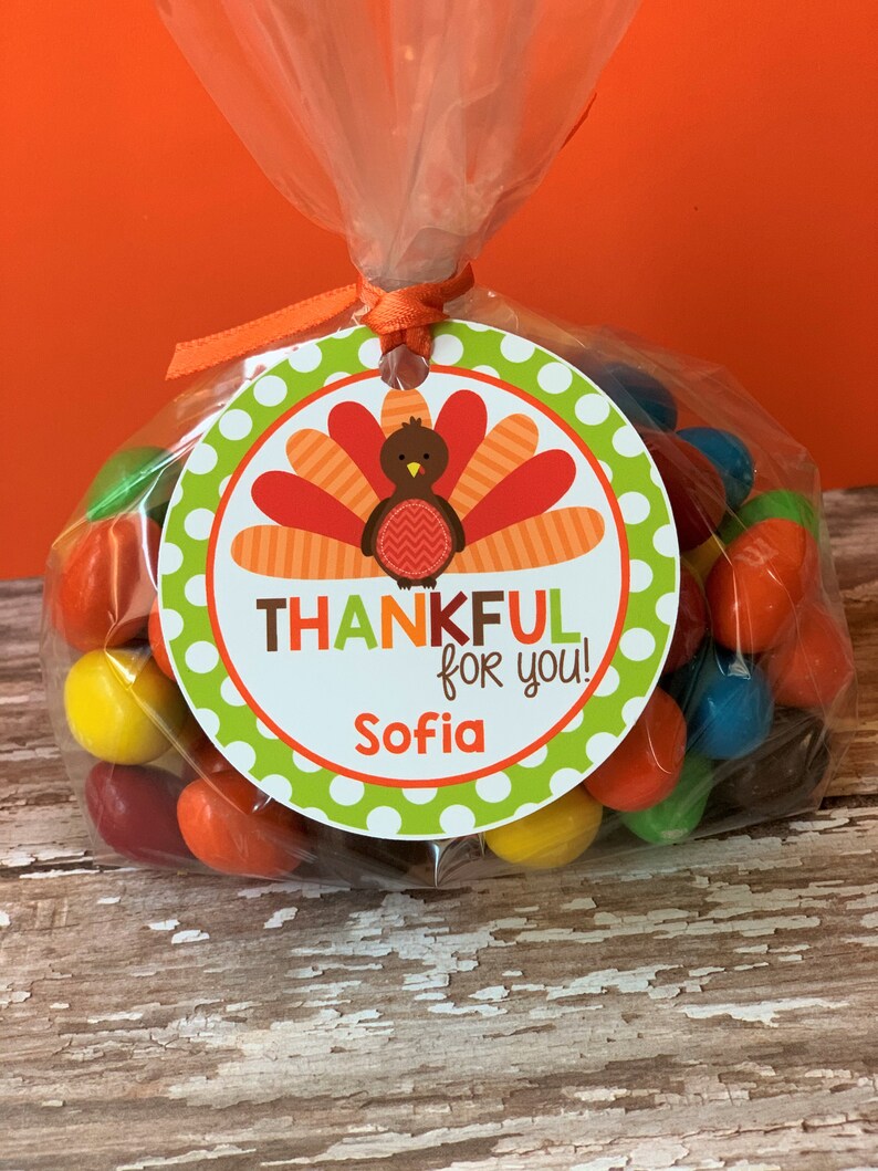 Op de afbeelding: Een ronde groene en witte tag met een kalkoenillustratie en de tekst "THANKFUL for you! Sofia". De tag is bevestigd aan een doorzichtige plastic zak gevuld met kleurrijke snoepjes.