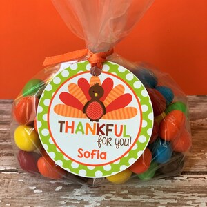 Op de afbeelding: Een groen en wit rond etiket met een Thanksgiving-kalkoenontwerp. Het etiket zegt "Thankful for you!" en "Sofia". Het etiket is bevestigd aan een doorzichtige plastic zak gevuld met kleurrijke snoepjes.