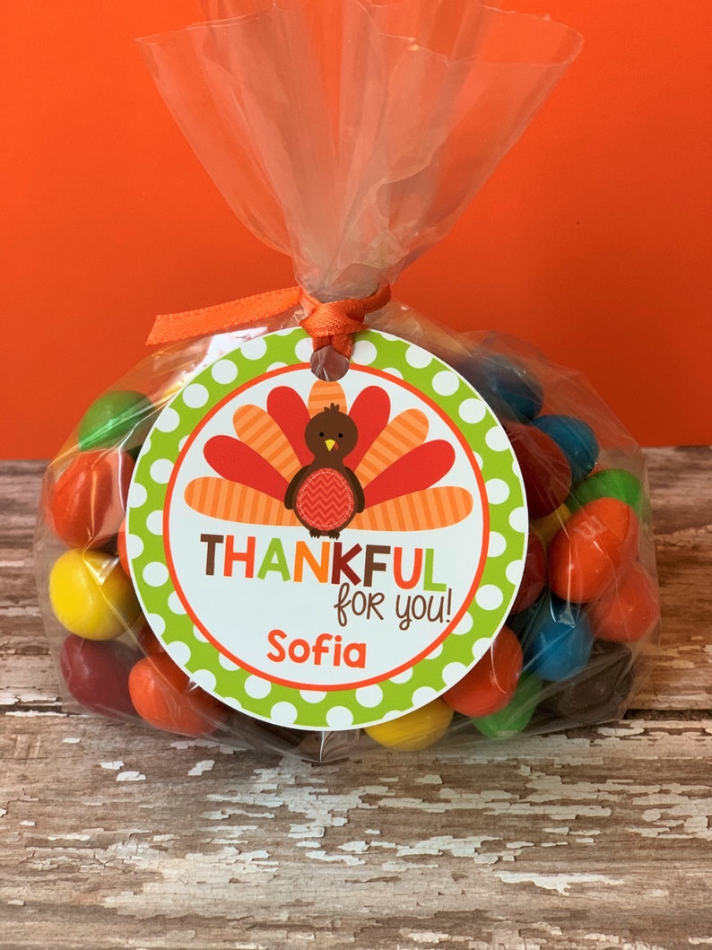Op de afbeelding: Een groen en wit rond etiket met een Thanksgiving-kalkoenontwerp. Het etiket zegt "Thankful for you!" en "Sofia". Het etiket is bevestigd aan een doorzichtige plastic zak gevuld met kleurrijke snoepjes.