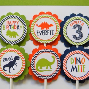 May include: Six green, orange and blue cupcake toppers with dinosaur designs. The toppers feature a green dinosaur, a red dinosaur, a blue dinosaur, the words "Happy Birthday", "Roar!" and "Dino Mite", and the name "Everett" with the number "3".