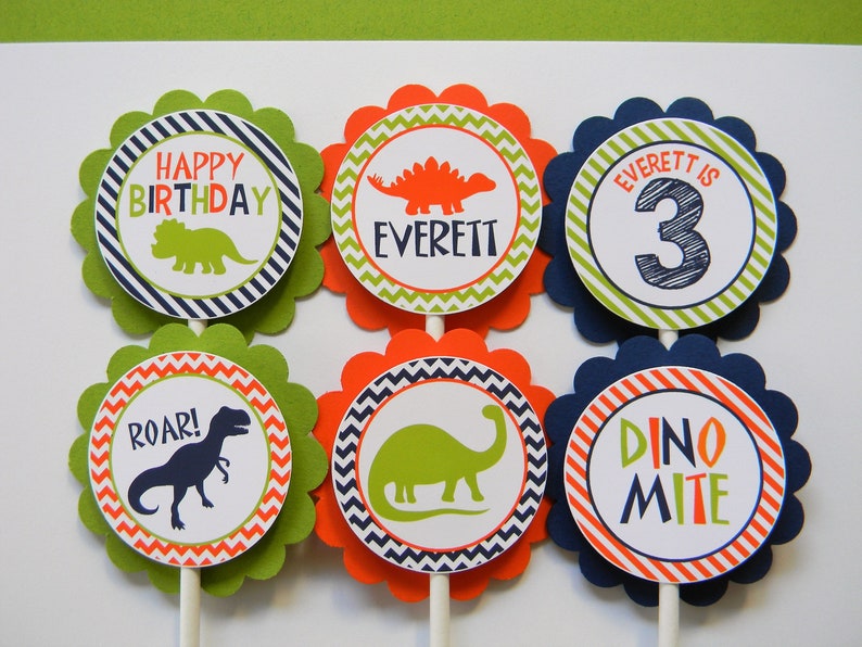 May include: Six green, orange and blue cupcake toppers with dinosaur designs. The toppers feature a green dinosaur, a red dinosaur, a blue dinosaur, the words "Happy Birthday", "Roar!" and "Dino Mite", and the name "Everett" with the number "3".
