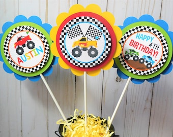 May include: Three monster truck themed centerpieces with a checkered flag design. The centerpieces feature a red monster truck, a yellow monster truck, and a green monster truck. One centerpiece says "Happy 4th Birthday!" Another centerpiece says "Austin".