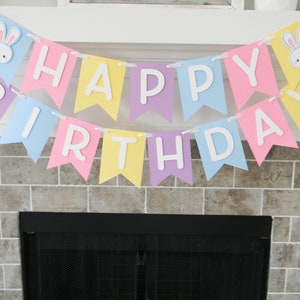 Puede incluir: Una pancarta de colores pastel con las palabras "Happy Birthday" en letras blancas. La pancarta tiene recortes de conejos en cada extremo.