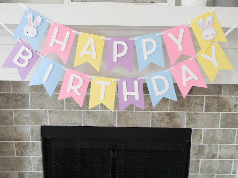 Puede incluir: Una pancarta de colores pastel con las palabras "Happy Birthday" en letras blancas. La pancarta tiene recortes de conejos en cada extremo.