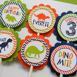 May include: Six green, orange and blue cupcake toppers with dinosaur designs. The toppers feature a dinosaur silhouette, the words "Happy Birthday", "Roar!" and "Dino-mite", and the name "Everett" with the number "3".