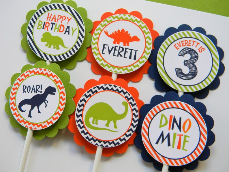 May include: Six green, orange and blue cupcake toppers with dinosaur designs. The toppers feature a dinosaur silhouette, the words "Happy Birthday", "Roar!" and "Dino-mite", and the name "Everett" with the number "3".