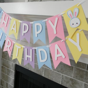 Puede incluir: Una pancarta de feliz cumplea&ntilde;os de colores pastel con letras blancas y recortes de conejos. La pancarta est&aacute; enhebrada en una cinta blanca y est&aacute; colgada sobre una chimenea.