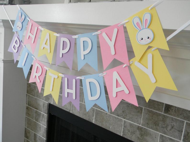 Puede incluir: Una pancarta de feliz cumplea&ntilde;os de colores pastel con letras blancas y recortes de conejos. La pancarta est&aacute; enhebrada en una cinta blanca y est&aacute; colgada sobre una chimenea.