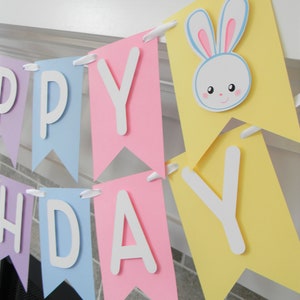 Puede incluir: Una pancarta de feliz cumplea&ntilde;os de colores pastel con un recorte de conejo blanco. La pancarta est&aacute; hecha de papel y tiene un borde festoneado. Las letras son blancas y el conejo es blanco con orejas rosas y ojos azules.