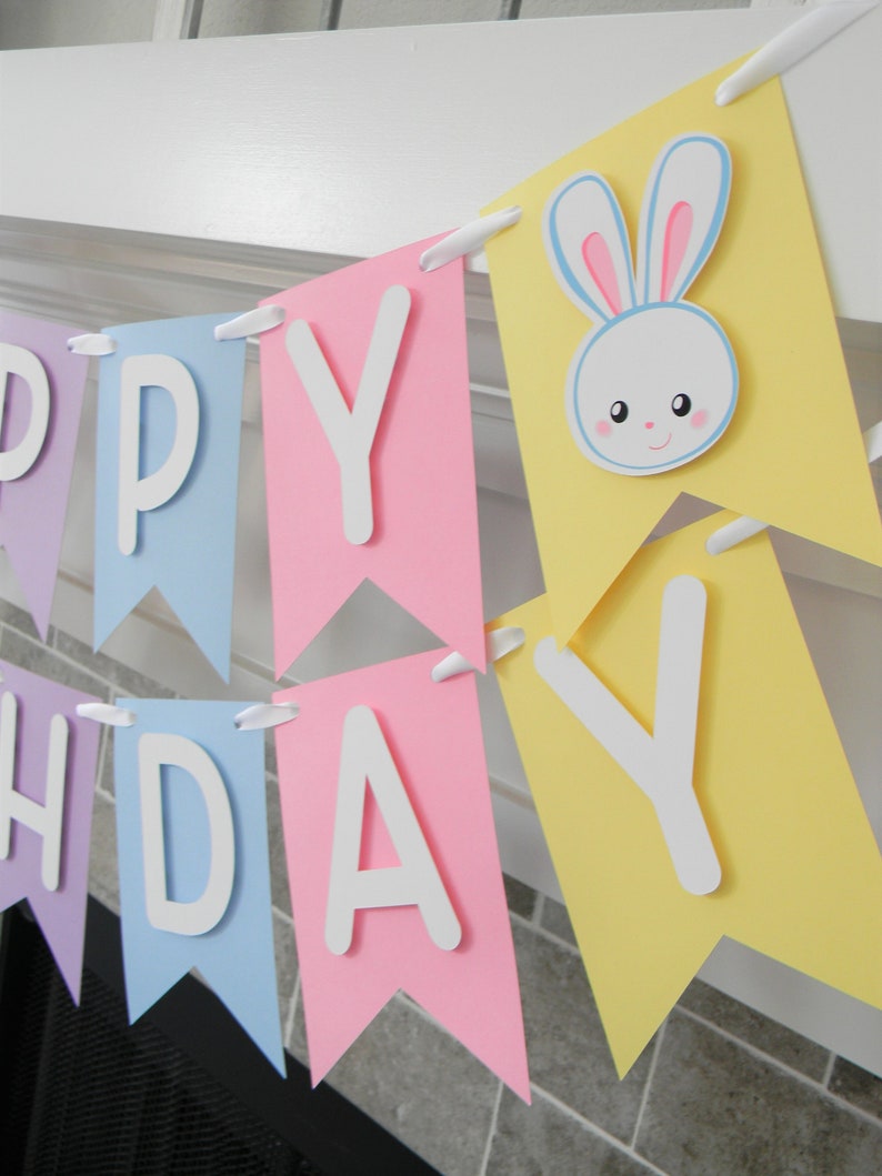 Puede incluir: Una pancarta de feliz cumplea&ntilde;os de colores pastel con un recorte de conejo blanco. La pancarta est&aacute; hecha de papel y tiene un borde festoneado. Las letras son blancas y el conejo es blanco con orejas rosas y ojos azules.