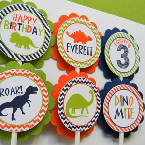 May include: Six green, orange and blue cupcake toppers with dinosaur designs. The toppers feature a green dinosaur, an orange dinosaur, a blue dinosaur, the words "Happy Birthday", "Roar!" and "Dino Mite", and the text "Everett is 3".