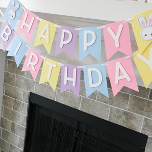 Puede incluir: Una pancarta de colores pastel con las palabras "Happy Birthday" en letras blancas. La pancarta est&aacute; decorada con recortes de conejos.