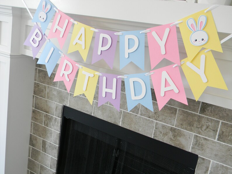 Puede incluir: Una pancarta de colores pastel con las palabras "Happy Birthday" en letras blancas. La pancarta est&aacute; decorada con recortes de conejos.