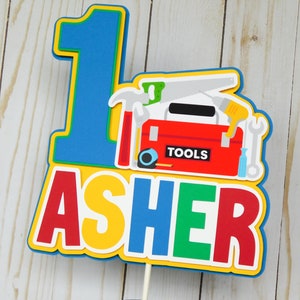 May include: A colorful cake topper with the number "1" and the name "Asher" in large letters. The number is blue, the name is yellow, red, green, and blue. The topper also features a red toolbox with various tools, including a hammer, saw, wrench, and screwdriver.