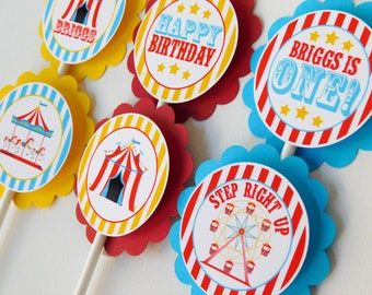 Könnte beinhalten: Eine Sammlung runder Papier-Cupcake-Topper auf weißen Stäbchen mit Jahrmarkt-Motiven und rot, gelb und blau gewellten Rändern. Die Grafiken zeigen ein Karussell, ein Zirkuszelt, ein Riesenrad sowie die Texte Happy Birthday, Briggs is One und Step Right Up.