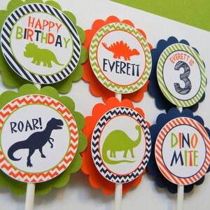May include: Six green, orange and blue cupcake toppers with dinosaur designs. The toppers feature a dinosaur silhouette, the words "Happy Birthday", "Roar!" and "Dino-mite", and the name "Everett" with the number "3".