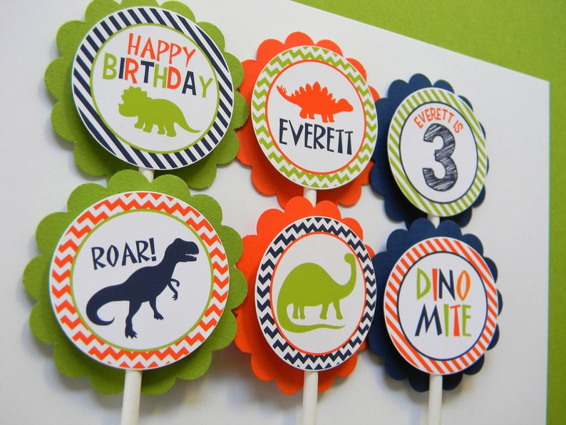 May include: Six green, orange and blue cupcake toppers with dinosaur designs. The toppers feature a dinosaur silhouette, the words "Happy Birthday", "Roar!" and "Dino-mite", and the name "Everett" with the number "3".